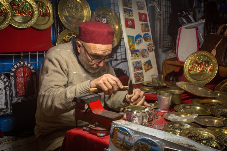 Sousse Tunisia - Dec 20 2023: A metal working craftsman wearing a fez hat uses a hammer at his tourist souvenir stall within the souk inthe ancient medina in Sousse, Tunisia. The medina is a UNESCO ; Shutterstock ID 2413844861; purchase_order: aljazeera ; job: ; client: ; other: