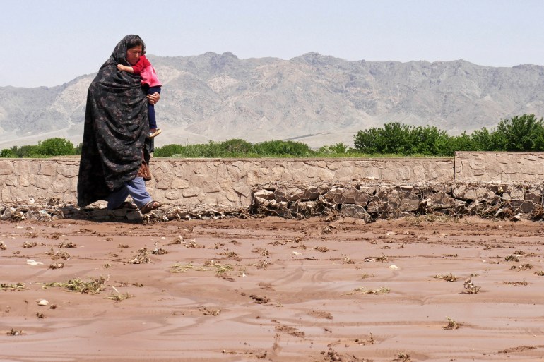 An Afghan woman carrying a child walks along a street covered in wet mud following flash floods in Herat on May 5, 2024. (Photo by Mohsen KARIMI / AFP)