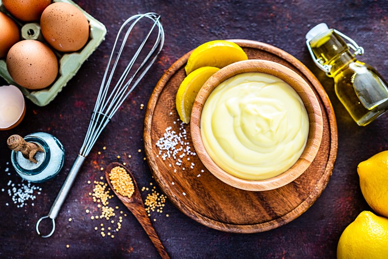 Food recipe: homemade mayonnaise in a wood bowl shot from above on dark rustic table. Ingredients for preparing homemade mayonnaise like olive oil, eggs, lemon and salt are around the prepared mayonnaise bowl. Predominant colors are brown and yellow. High resolution 42Mp studio digital capture taken with SONY A7rII and Zeiss Batis 40mm F2.0 CF lens