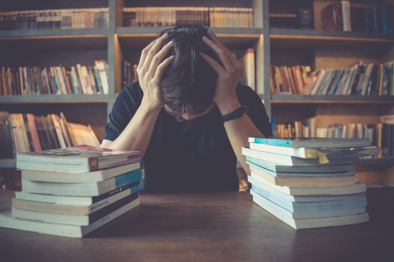 Stress and tired man under mental pressure while reading book preparing examination in library. Businessman stressed overworked studying on desk in library. Headache, migraine, education concept.