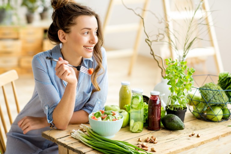 Young and happy woman eating healthy salad sitting on the table with green fresh ingredients indoors