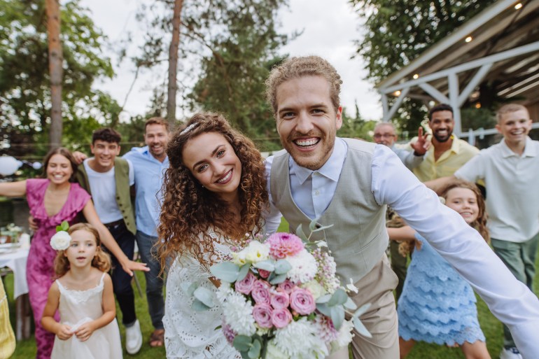 Family portrait from a small garden wedding. The bride and groom standing in front their parents and friends in formal attire standing in the garden and pose for a photo.