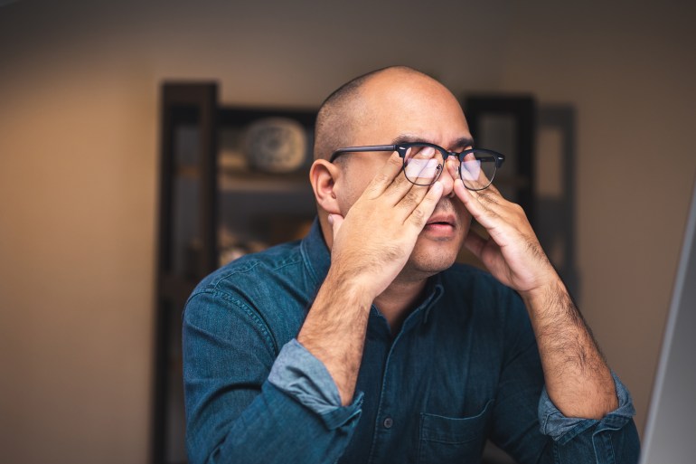 A young businessman works with computer late at night in a dimly lit room. He is tired and stressed, needs rest. Take off glasses and rubbing his eyes