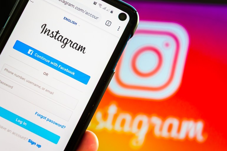 Closeup Of A Hand Holding Mobile Smart Cell Phone Device With Instagram Social Network Log In Screen Over a Notebook With Instagram Web Image On Display