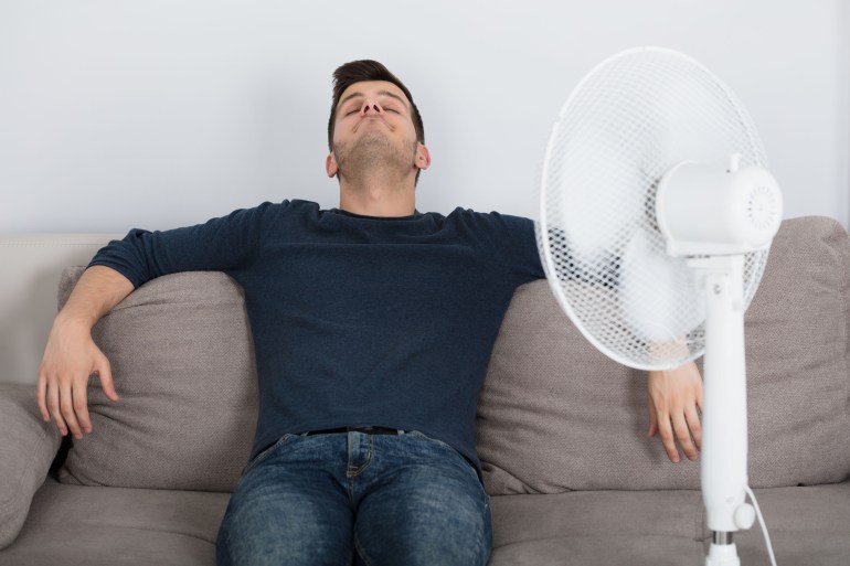 Young Man Sitting On Couch Cooling Off With Fan During Hot Weather At Home; Shutterstock ID 734181577; purchase_order: ajanet; job: ; client: ; other: