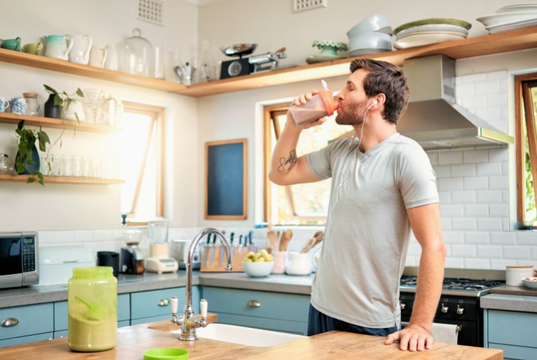 One fit young caucasian man drinking bottle of chocolate whey protein shake for energy for training workout while wearing earphones in a kitchen at home. Guy having sports supplement for muscle gain and dieting with weightloss meal replacement