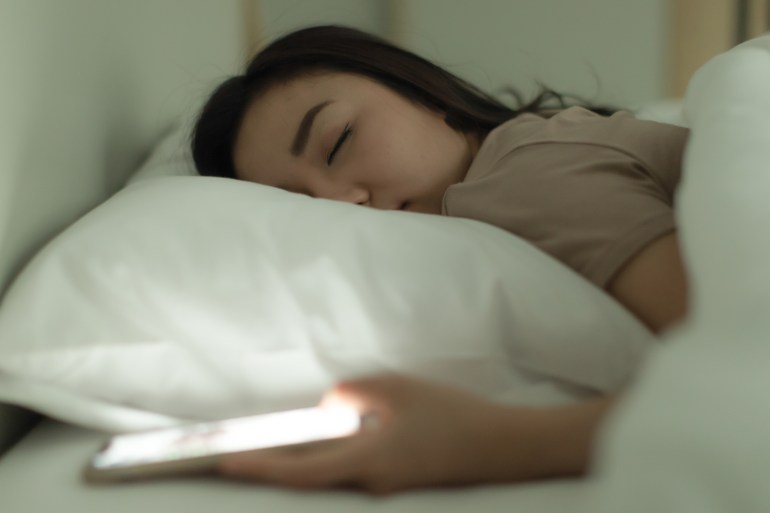 Cute young Asian woman laying on the bed while holding the switch on an open light smartphone in hand and asleep, nap with tired and deep sleep. Social media addict concept; Shutterstock ID 2176356905; purchase_order: aljazeera ; job: ; client: ; other: